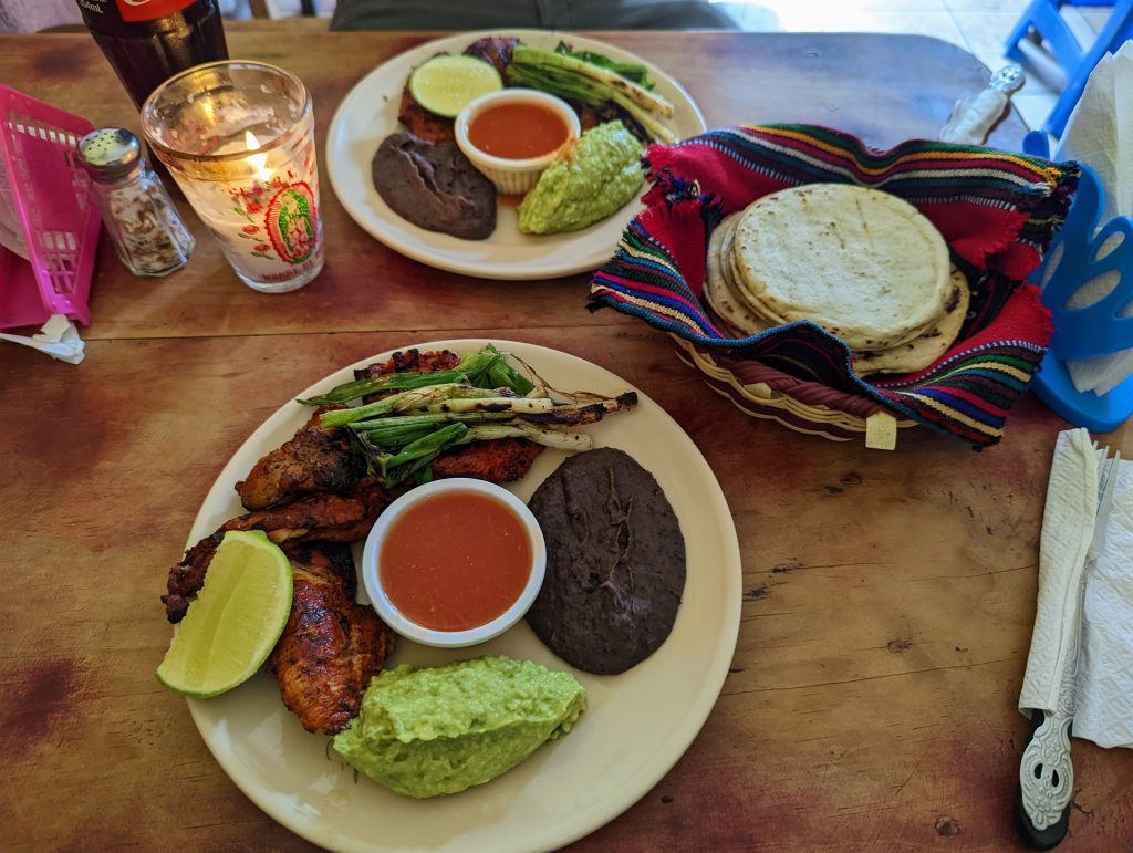 Two plates on a wood table containing guacamole, beans, pork, green onions and lime. 