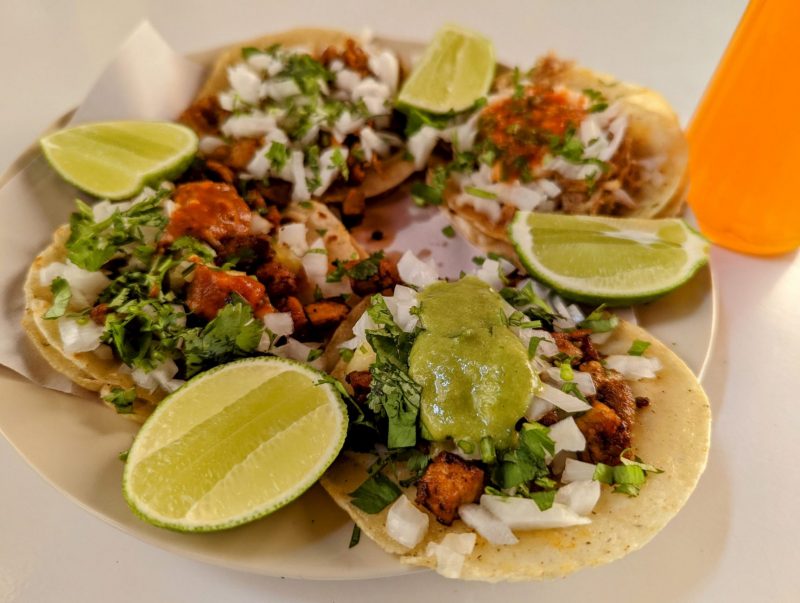 Four Tacos on a white plate. Four slices of lime are on the plate. 