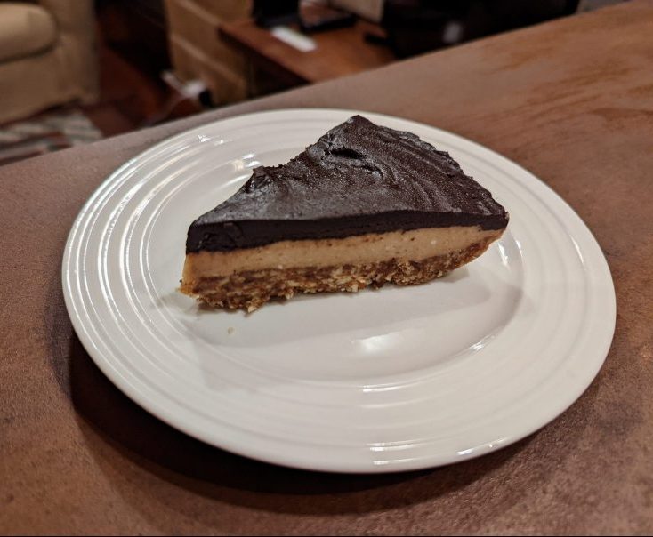 Slice of dessert on a round white place. The dessert has a dark chocolate top layer, a neutral middle layer and a nutty bottom layer. 