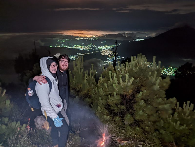 Man and women standing in the bushes with a fire below. It is night time and you can see the lights from the city in the distance. 