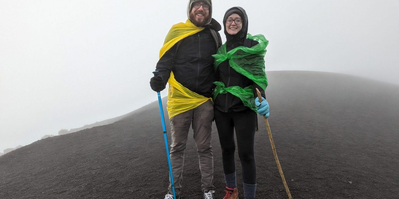 Woman and man standing on top of a volcano. The ground is black and the sky white.