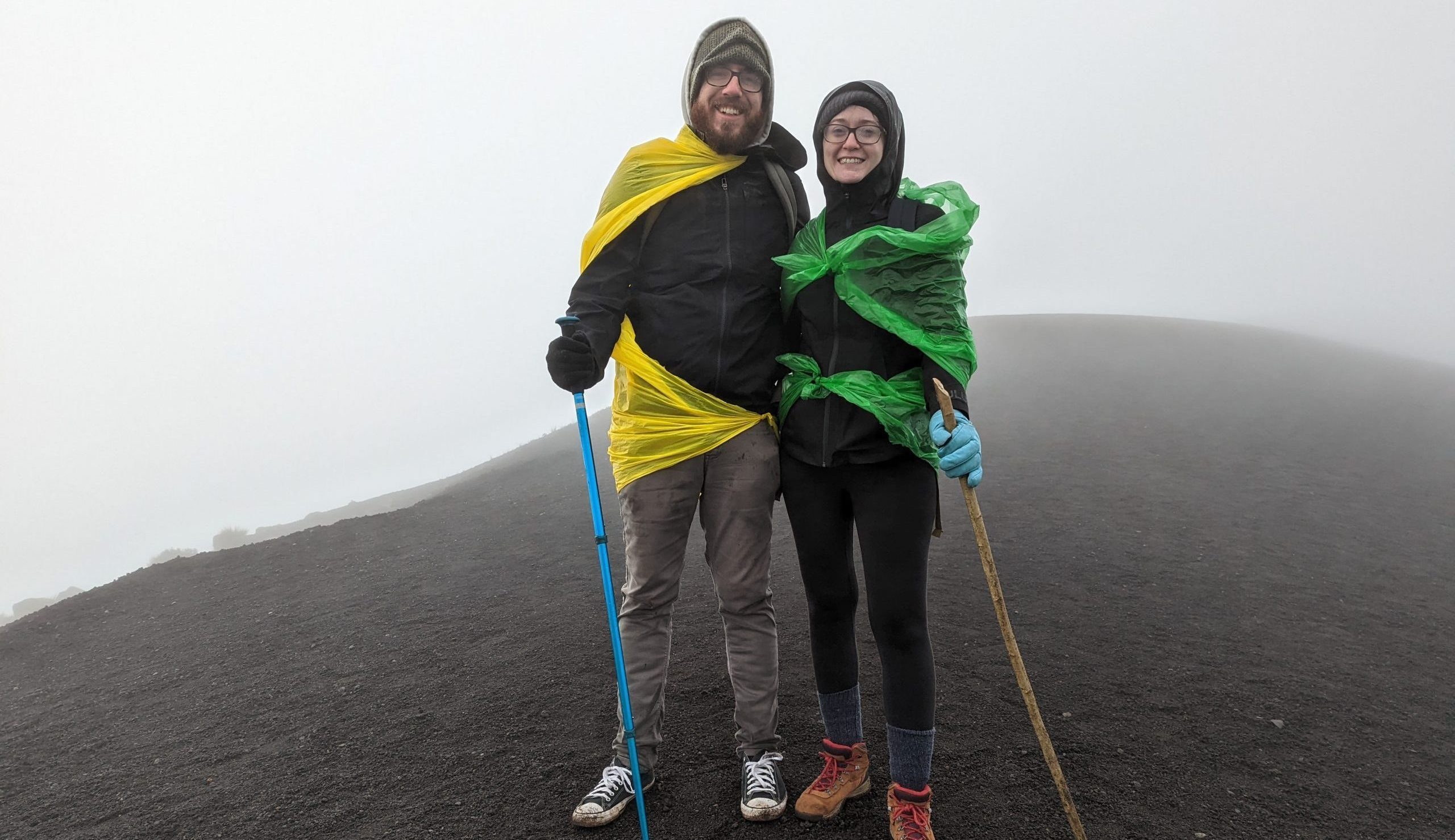 Woman and man standing on top of a volcano. The ground is black and the sky white.