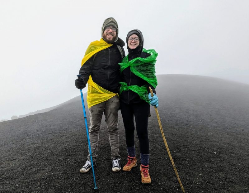 Jessica and Jonathan on the top of the volcano. They are wearing warm clothing and holding walking sticks. 