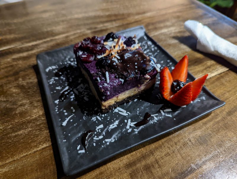 Purple square dessert with strawberry and blueberries, on a black plate. 