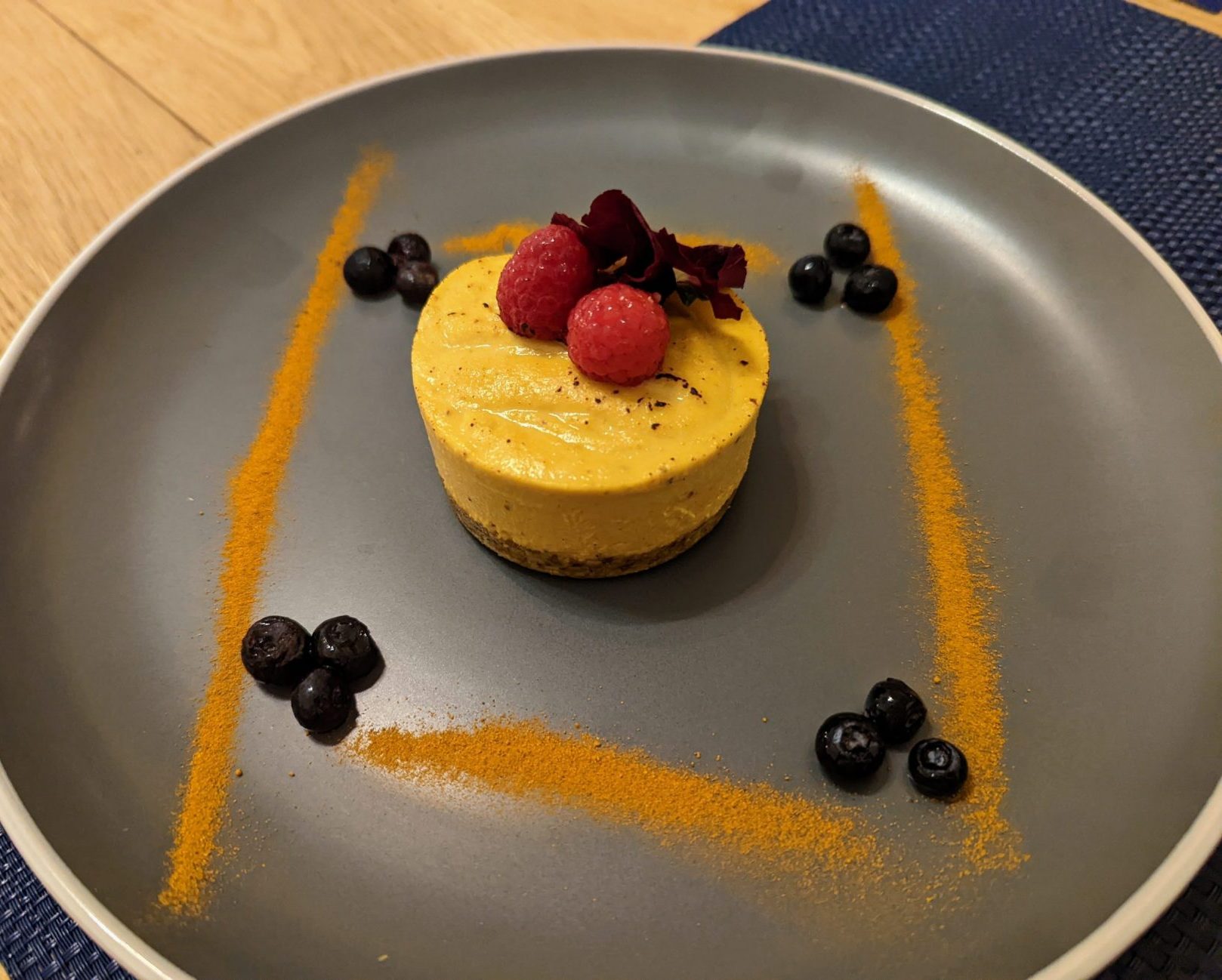 Picture of a small round yellow dessert, topped with two raspberries and surrounded by blueberries and spices. 