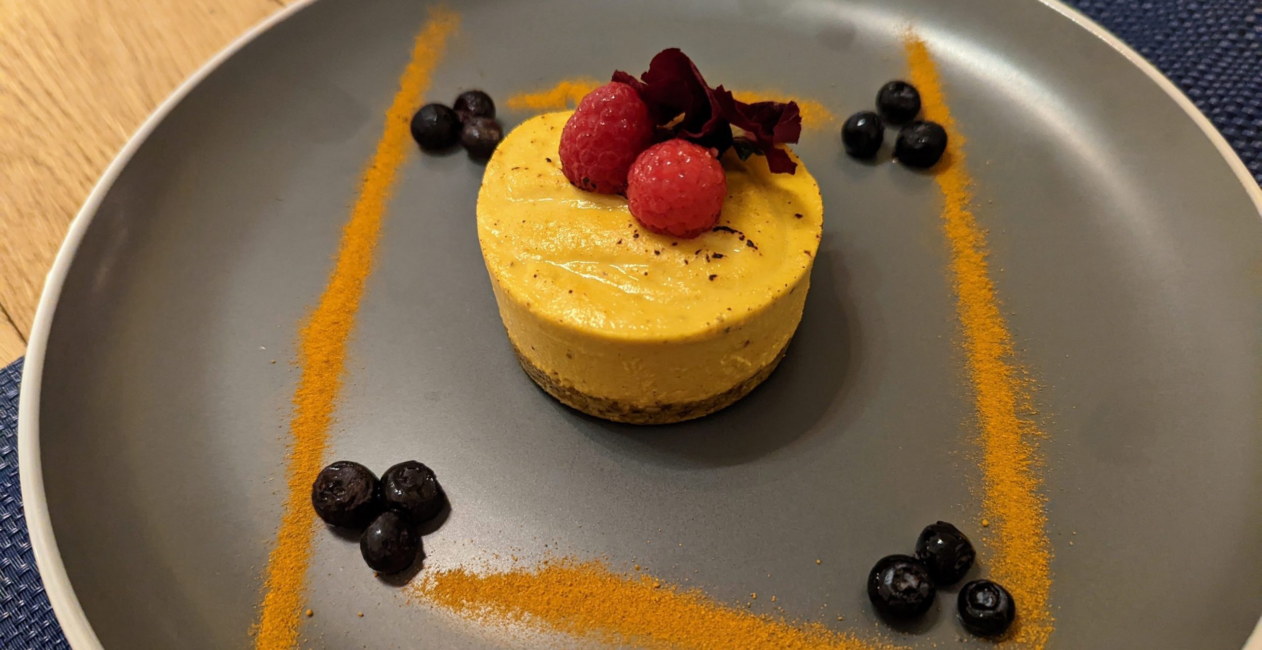 A yellow round dessert, topped with two raspberries and surrounded by blueberries and spices.