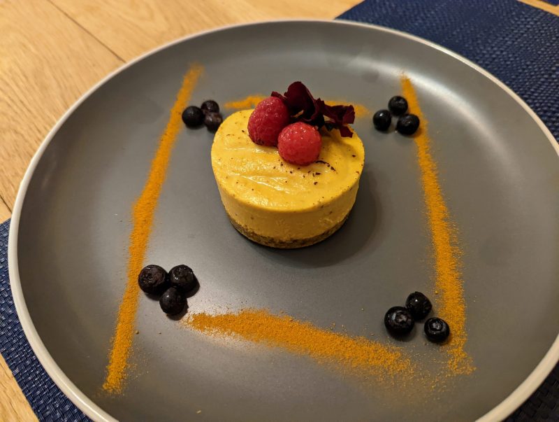 Yellow round dessert, topped with two raspberries and surrounded by blueberries and spices. 