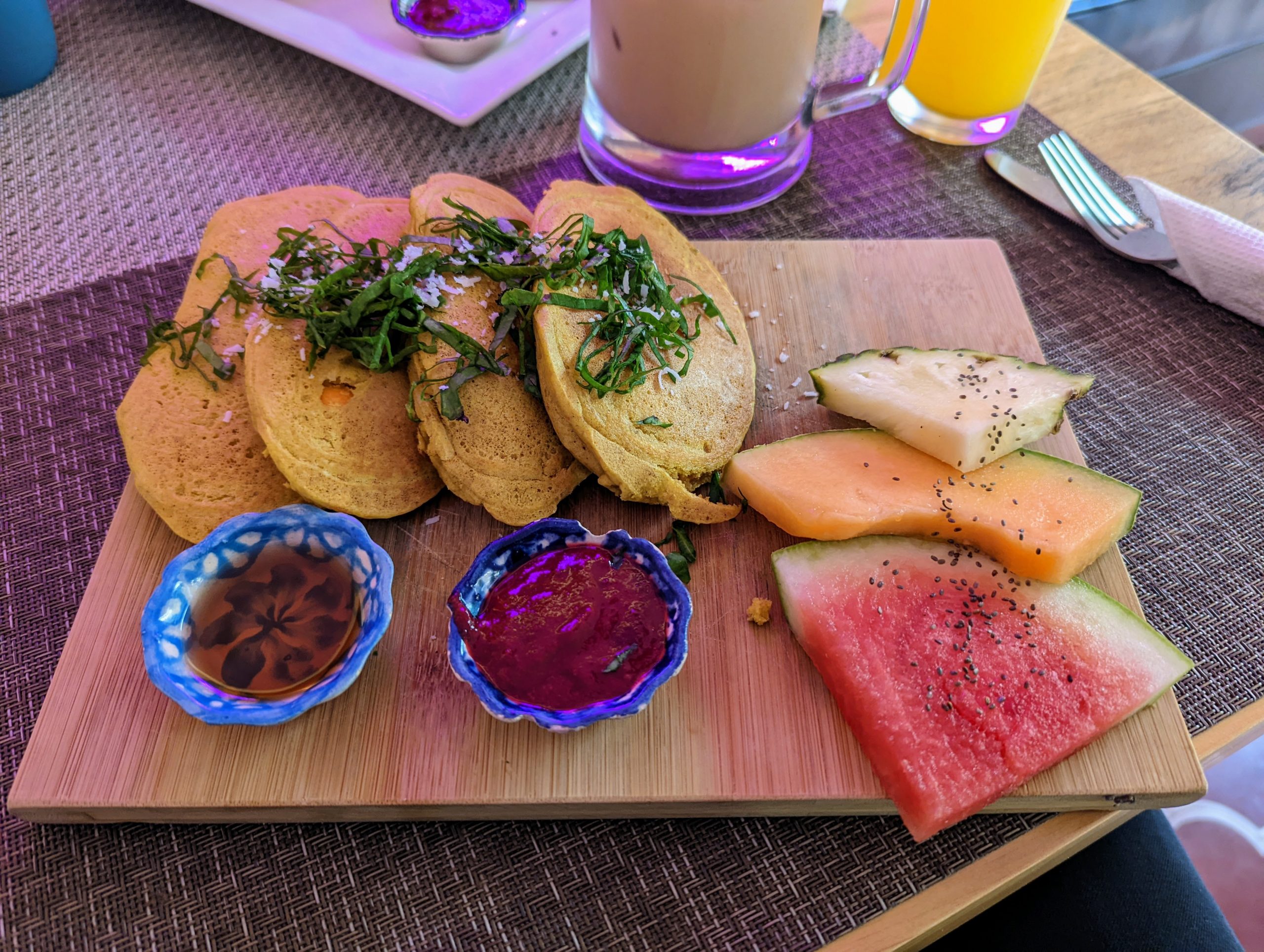 A cutting board with four pancakes, melon slices and two small bowls containing condiments on top. 