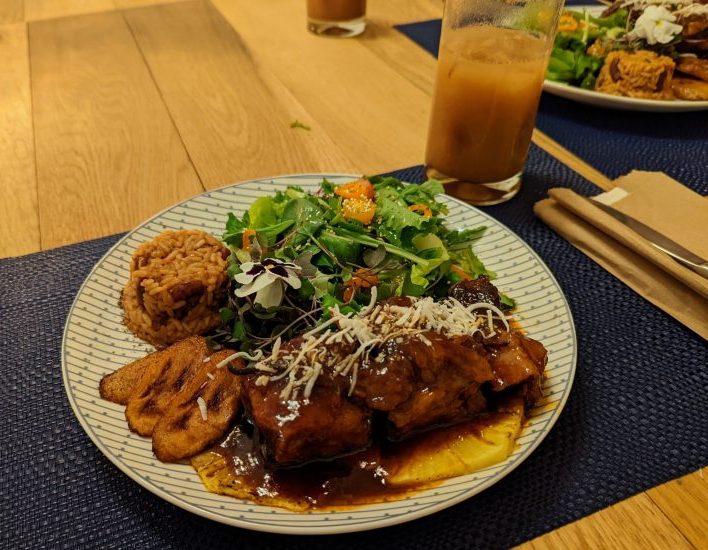 Pork ribs coveredd in a brown sauce, on top of a pinapple slice. Fried plantain slices, a small portion of rice and a green salad are also on the plate.