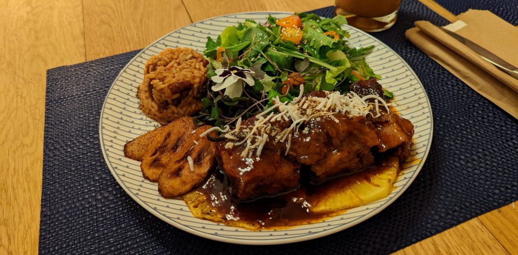 Pork ribs coveredd in a brown sauce, on top of a pinapple slice. Fried plantain slices, a small portion of rice and a green salad are also on the plate.