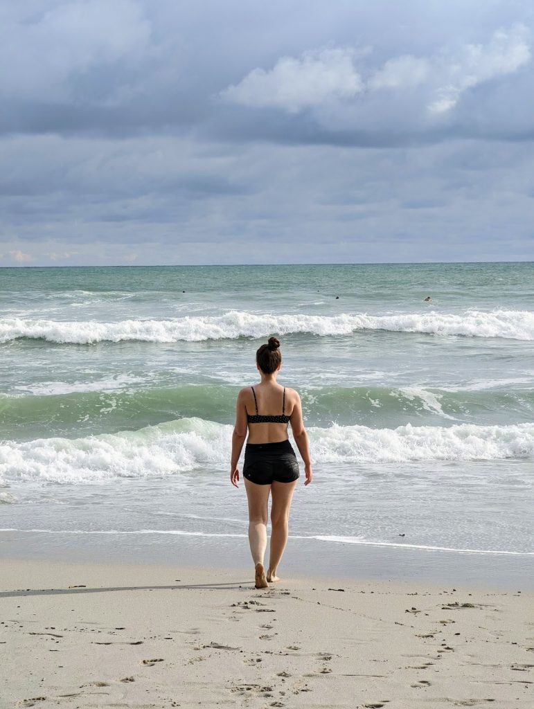 A woman wearing black shorts and a black swim top is walking towards the ocean. The water is green and blue and waves can be seen,.