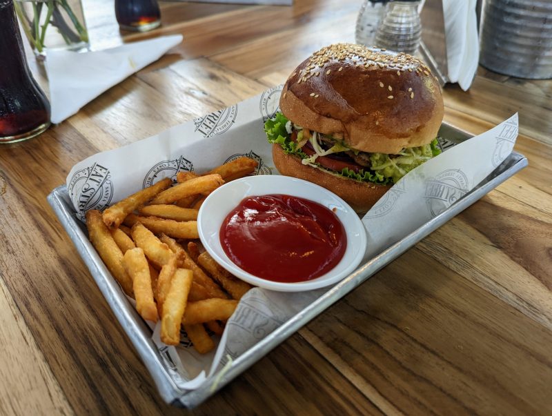 Tuna Burger with fries, a small white bowl containing ketchup, all in a metal try on a wooden table.table.