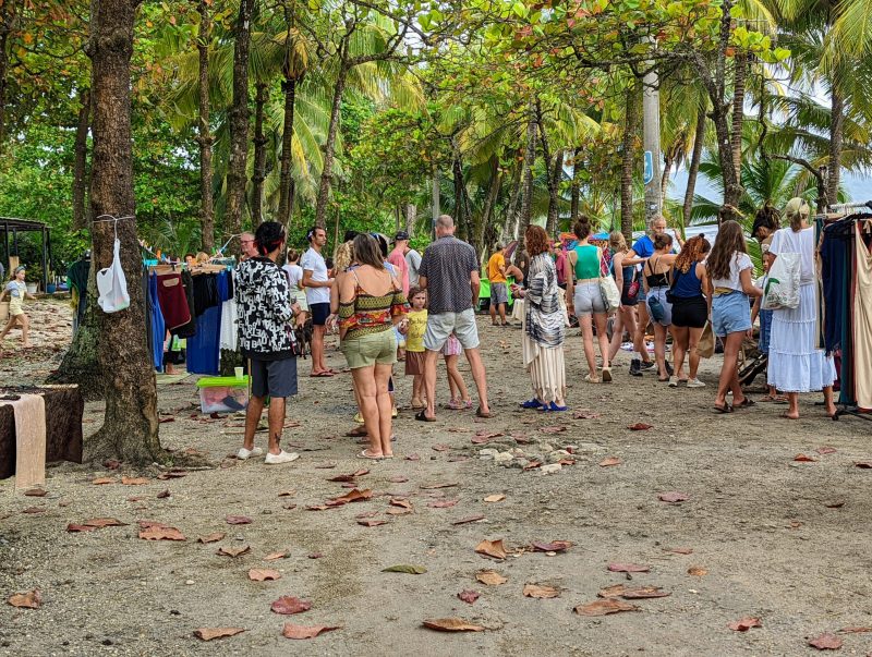 People talking and visiting different merchants looking at products. The ground is covered in sand and there are trees around the people.