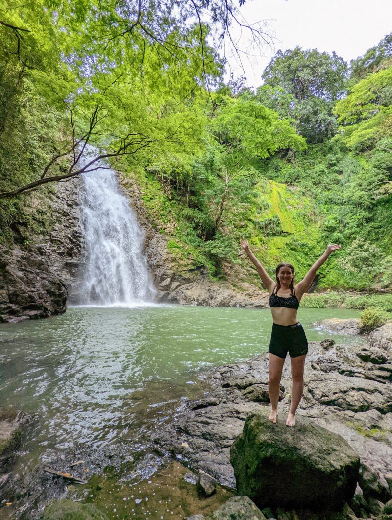 Woman standing on a rock with her arms raised above her head. Behind her is a waterfall. 